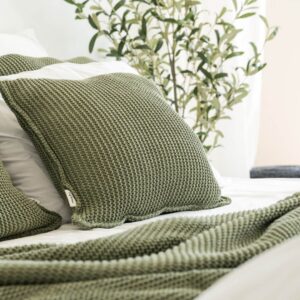 copricuscino lavorato a maglia con filato a trama grossa color verde oliva di misura 50x50 utilizzato su un letto con altri cuscini e coperte