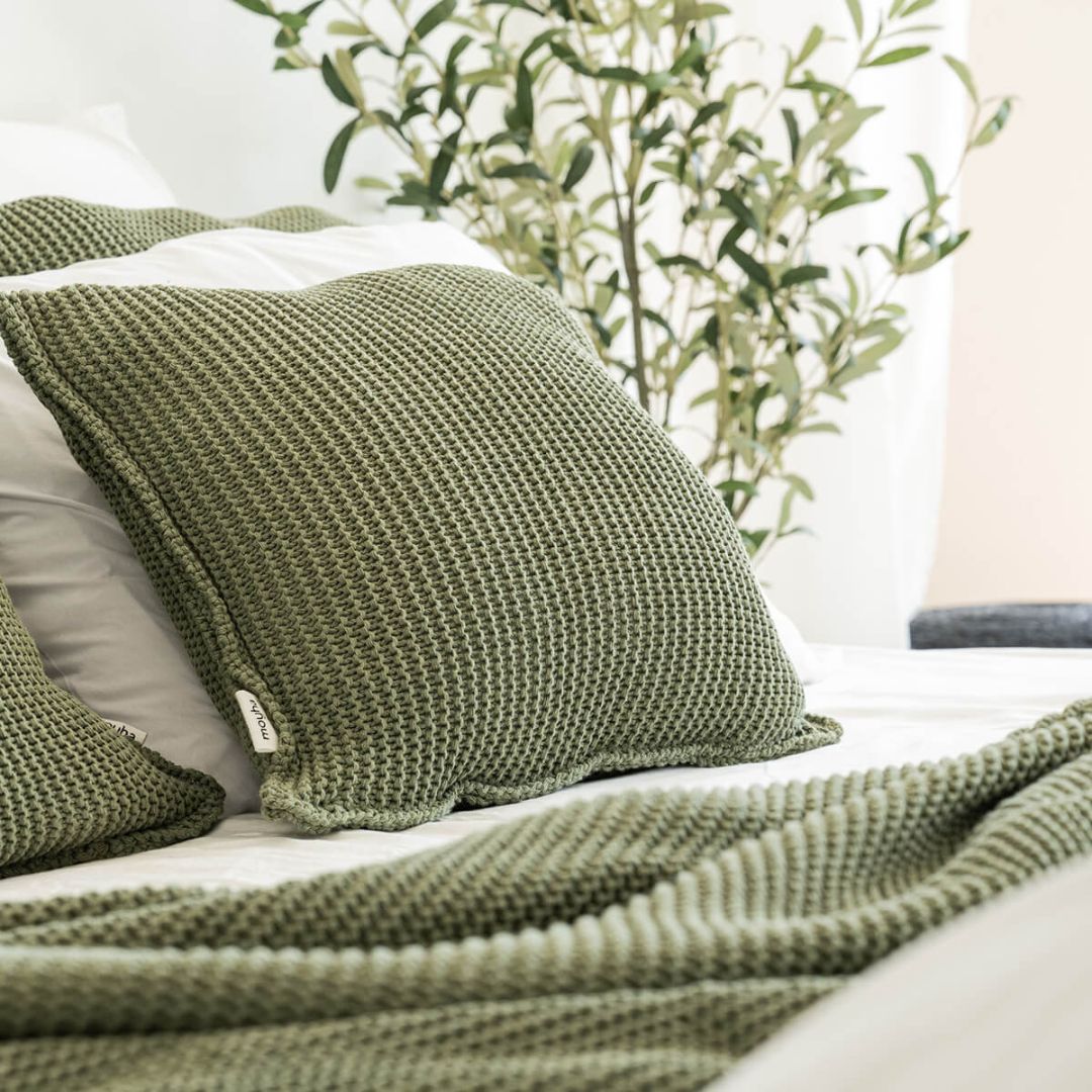 copricuscino lavorato a maglia con filato a trama grossa color verde oliva di misura 50x50 utilizzato su un letto con altri cuscini e coperte