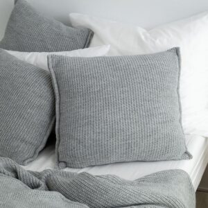 copricuscino lavorato a maglia con filato a trama grossa color grigio chiaro di misura 60x60 utilizzato su un letto con altri cuscini e coperte
