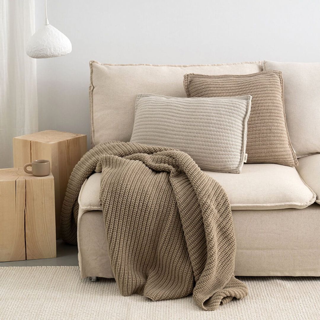 salotto con divano beige chiaro impresiosito da cuscini e coperta in lana lavorata a maglia di color beige e taupe
